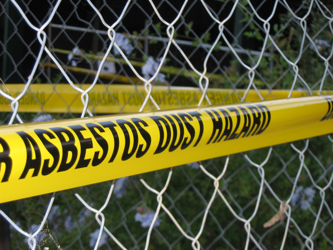 Caution tape around chainlink fence says asbestos dust hazard